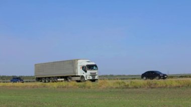 Güneşli bir havada beyaz bir araba otoban boyunca sürer. Otoyolda kamyon var..