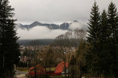 Bucegi Dağları (Romanya 'nın orta kesimindeki Karpat Dağları' nın Güney Karpatlar grubunun bir parçası) soğuk ve bulutlu bir kış sabahı Busteni kasabasından görüldüğü gibi arka planda yer alır..