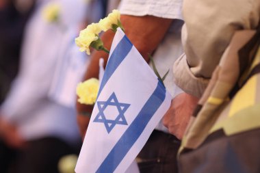 Ekim 2023 saldırılarının anısına düzenlenen törende İsrail bayrağı ve çiçek tutan bir adamın elleri ile ilgili detaylar.