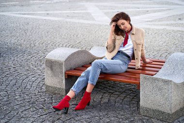 Güzel, genç, kızıl saçlı bir kadın. Oyuncak ceketli, kot pantolonlu, kırmızı yakalı bir kadın. Kentteki bir bankta oturuyor. Şehir hayalleri.