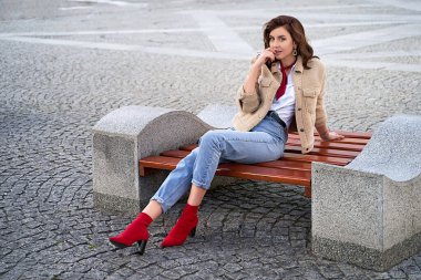 Güzel, genç, kızıl saçlı bir kadın. Oyuncak ceketli, kot pantolonlu, kırmızı yakalı bir kadın. Kentteki bir bankta oturuyor. Şehir hayalleri.