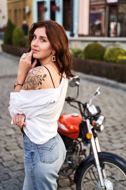 Kot pantolonlu ve beyaz gömlekli genç bir kadın sokakta motosikletin yanında poz veriyor.