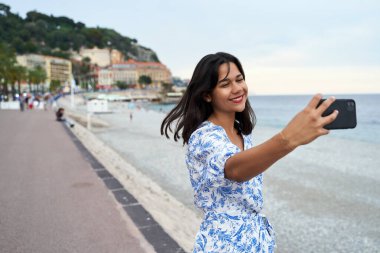 Güzel, gülümseyen genç bir kadın, Fransa 'nın Nice kentindeki Gezinti Güvertesi' nde akıllı telefonuyla selfie çekiyor.