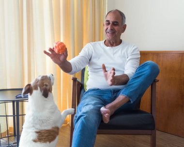 63 yaşında, rahat bir koltukta oturan, gülümseyen ve içeride Jack Russell teriyeri köpeğiyle oynayan bir Türk. Neşeli an, evcil hayvanla sahibinin sıcak bir ev ortamındaki güçlü bağını yakalar..