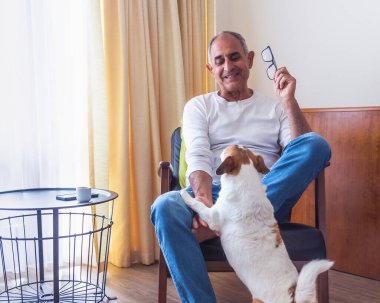 Oturduğu koltukta oturan ve içeride Jack Russell teriyeri köpeğiyle oynayan Türk kökenli gülümseyen kıdemli adam. Samimi an sıcaklığı, dostluğu ve rahat bir aile ortamında günlük yaşamı gösteriyor..