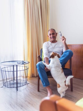 Bir koltukta oturan ve enerjik Jack Russell teriyer köpeği ile içeride oynayan Türk kökenli gülümseyen kıdemli adam. Samimi an sıcaklığı, dostluğu ve rahat bir aile ortamında günlük yaşamı gösteriyor..