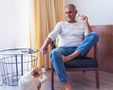 Gözlüklü yaşlı bir adam koltukta oturuyor ve enerjik Jack Russell teriyeri köpeğiyle içeride oynuyor. Samimi an sıcaklığı, dostluğu ve rahat bir aile ortamında günlük yaşamı gösteriyor..