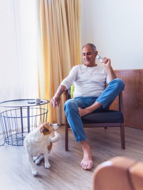 Bir koltukta oturan ve enerjik Jack Russell teriyer köpeği ile içeride oynayan Türk kökenli gülümseyen kıdemli adam. Samimi an sıcaklığı, dostluğu ve rahat bir aile ortamında günlük yaşamı gösteriyor..