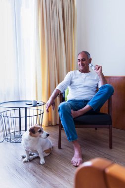 Düşünceli yaşlı bir adam, sadık Jack Russell teriyerini ayaklarının dibinde seyrediyor. İçerideki sakin sahne, insan ve evcil hayvan arasındaki basit ve anlamlı bağlantıyı yakalar..