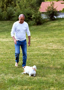 Sıcak bir yaz gününde yeşil çimlerde Jack Russell Terrier 'ıyla eğlenen neşeli son sınıf öğrencisi. Adam onu cesaretlendirirken köpek etrafta koşuşturur. Uzaktaki ev..