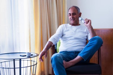 Düşünceli yaşlı bir adam, rahat bir odada tek başına oturuyor, elinde okuma gözlüğü var. Yakın bir masadaki kişisel eşyalarla sakin bir yansıma anı..