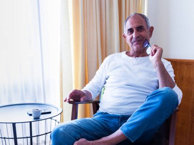 Yaşlı adam güneşli bir odada sessizce oturuyor, gözlüklerini tutuyor ve düşüncelere dalıyor. Bir cep telefonu ve küçük bir kahve fincanı yan masada dinleniyor..
