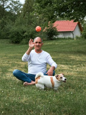 Sakin, yalın ayak, yaşlı bir adam yeşil çimlerin üzerinde oturuyor ve Jack Russell Terrier 'ı için bir top fırlatıyor, o da tam gaz koşuya çıkıyor. Köpek, arka planda bir evle güneşli bir bahçede heyecan ve enerji yayıyor..