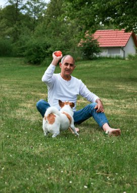 Kıdemli Ma çıplak ayaklı yaşlı bir adam yeşil bir yaz çimenliğinde oturur ve Jack Russell Terrier 'ının önünde bir top tutar. Köpek oyuncağa doğru hevesle uzanır ve neşeli bir etkileşim anı yaratır. Arka planda bir ev görünüyor. Tam oldu.