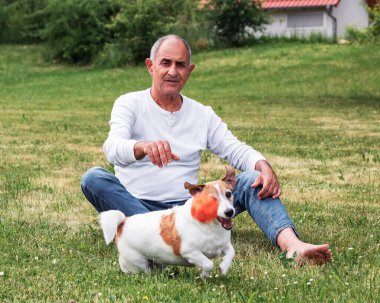 Sağlıklı yaşlı bir adamın enerjik teriyerine top yakalamayı öğretmesine odaklan. Hareket ve neşe dolu bir yaz günü. Arka planda rahat bir ev ve yeşil çimenler görünüyor..