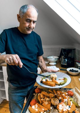 Kıdemli adam elinde lezzetli kızarmış sebzeler, karides, sarımsak ve peynir tabağı tutuyor. Fotoğraf özgüveni, kendine yeterliliği ve dengeli bir diyete olan bağlılığı yansıtıyor. Emeklilikte sağlıklı bir yaşam tarzının ideal görüntüsü.