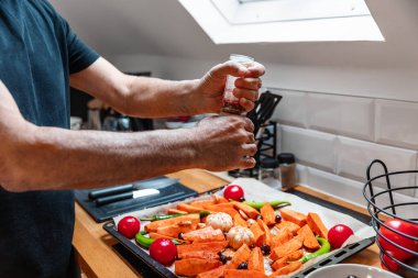 Modern bir tavan arası mutfağındaki taze sebze tepsisinin üzerinde biber öğütücü kullanan yaşlı erkek elleri ve tavan penceresinden gelen doğal ışık. Tatlı patates, domates, sarımsak, yeşil biber, zeytin ve zencefil parçaları hazır.