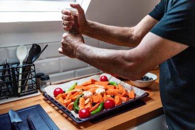 Modern bir tavan arasında gün ışığıyla dolu sebze çeşnili yaşlı ellerin detaylı bir çekimi. Tepside tatlı patates, domates, zeytin, sarımsak, yeşil biber ve zencefil gibi canlı malzemeler var. Sahne dikkatli beslenmeyi öneriyor.