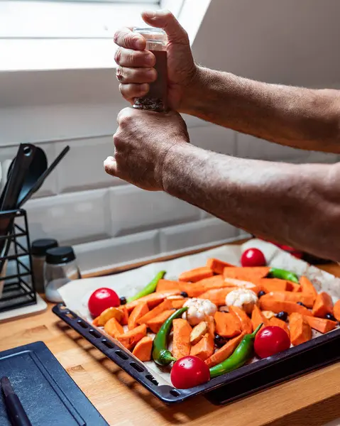 Modern bir minimalist tavan arası mutfağında, tepside renkli sebze karışımı pişiren yaşlı erkeklerin yakın çekim görüntüleri. Yemekler arasında domates, tatlı patates, sarımsak, zeytin, yeşil biber ve zencefil yer alıyor. Pencereden gelen doğal ışık