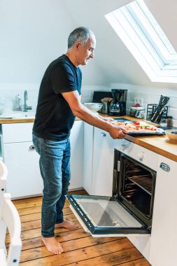 Son sınıf öğrencisi, çeşitli sebzelerle dolu bir tepsiyi modern tavan arası mutfağında minimalist tasarımı olan açık bir fırına koyar. Mekan, yakındaki bir pencereden gelen doğal ışıkla dolu. Fotoğraf ev yemeklerini ve bağımsızlıklarını gösteriyor.
