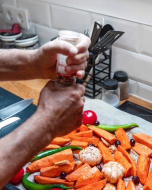 Minimalist, modern bir mutfakta sağlıklı bir sebze yemeği hazırlayan yaşlı erkeklerin yakın çekim görüntüsü. Tatlı patates, sarımsak, domates, dolmalık biber, zeytin ve zencefil gibi rengarenk malzemelerle dolu bir tepsi öğütülüyor.