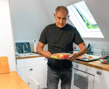 Parlak bir tavan arası mutfağında duran yaşlı adam elinde taze kavrulmuş sebzelerle dolu bir tabak tutuyor. Seyirciyle yüz yüze geldiğinde, sağlıklı bir ev yemeği ya da akşam yemeğinin tadını çıkarmaya hazırlanıyor. Beyaz minimalist mutfak pencereden gün ışığıyla dolu.