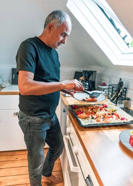 Parlak bir minimalist tavan arası mutfağında tabağa taze kızarmış sebze servis eden son sınıf bir adamın profili. Gün ışığı pencereden içeri giriyor, rahat yeri aydınlatıyor. Görüntü bağımsız yaşama, sağlıklı yeme alışkanlıklarına dikkat çekiyor, vejetaryen