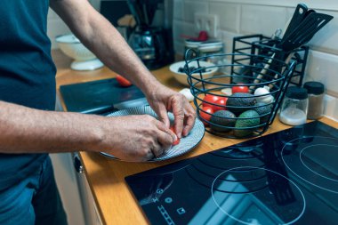 Son sınıf öğrencilerinin parlak, modern bir mutfakta büyük beyaz bir tabakta dilimlenmiş domatesleri sıralarken tepeden tırnağa görüntüsü. Mutfak masasının yanında içi taze domates, avokado ve sarımsak dolu bir sepet var. Sahnede sağlıklı beslenme, vejetaryen