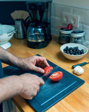 Son sınıf öğrencisinin, minimalist bir mutfakta tahta kesme tahtasıyla taze bir domatesi dilimlerken çekilmiş yakın çekimi. Tezgahın yanında bir kase yeşil zeytin duruyor. Sahne sağlıklı yeme alışkanlıklarını, kendi kendini hazırlayan vejetaryen yemeklerini ve sakinliğini yansıtıyor.