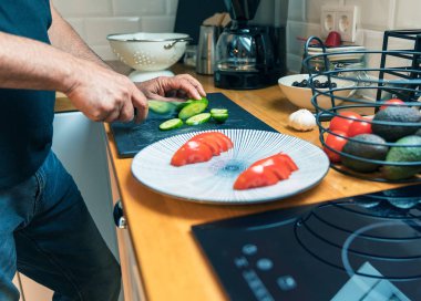 Parlak, modern bir mutfağın ahşap kesme tahtasının üzerinde taze yeşil bir salatalığı kesen erkeklerin yan görüntüsü. Kesme tahtasının yanında dilimlenmiş domateslerle dolu büyük beyaz bir tabak ve yanında da taze domates, avokado ve sarımsak dolu bir sepet var.