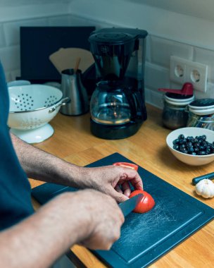 Son sınıf öğrencilerinin parlak, modern bir mutfakta ahşap bir kesme tahtasıyla taze bir domatesin doğrandığı tepeden tırnağa görüntüsü. Tezgahın yanında bir kase dolusu siyah zeytin ve sarımsak var. Sahne sağlıklı yemek ve taze malzemelere odaklanıyor.