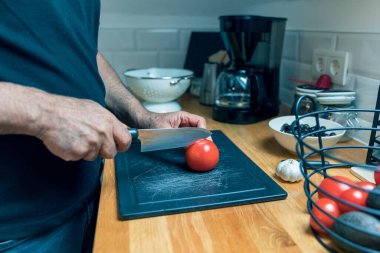Son sınıf öğrencilerinin parlak minimalist bir mutfakta ahşap bir kesme tahtasının üzerinde taze bir domatesi dilimlerken yan görüntüsü. Fotoğraf yemek pişirme sürecini yandan yakındaki avokado ve sarımsak içeren bir sepet sebzeyi göstererek kaydediyor. Doğal gün ışığı