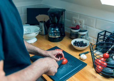 Son sınıf öğrencilerinin parlak, modern bir mutfakta ahşap bir kesme tahtasıyla taze domates kestikleri yakın plan çekimler. Büyük bir tabak sağlıklı bir salata için hazır. Yakınlarda olgun domatesler, avokado ve sarımsak var. Fotoğraf sağlıklı beslenmeyi ön plana çıkarıyor.