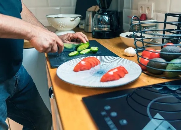 Parlak, modern bir mutfağın ahşap kesme tahtasının üzerinde taze yeşil bir salatalığı kesen erkeklerin yan görüntüsü. Kesme tahtasının yanında dilimlenmiş domateslerle dolu büyük beyaz bir tabak ve yanında da taze domates, avokado ve sarımsak dolu bir sepet var.