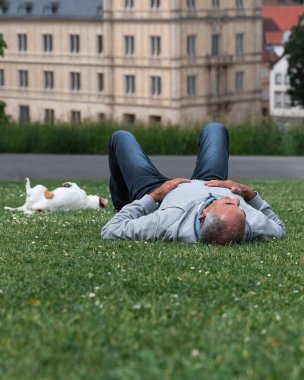 Hofgarten, Coburg 'da yeşil çimlerin üzerinde yatan kıdemli adamla Jack Russell terrier' i arasındaki bağlantı çok dokunaklı. Tema: Yaşlılıkta evcil hayvan arkadaşlık, güven ve duygusal bağ.