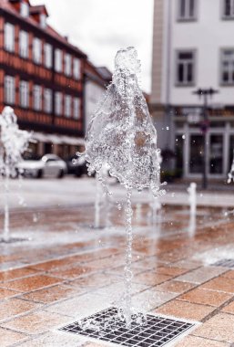 Kuru fıskiye fıskiyeleri Albertplatz Coburg Bavyera 'daki asfalttan su püskürtüyor. Arka planda bulanık şehir manzarası olan su sıçratmaya odaklanıyor.