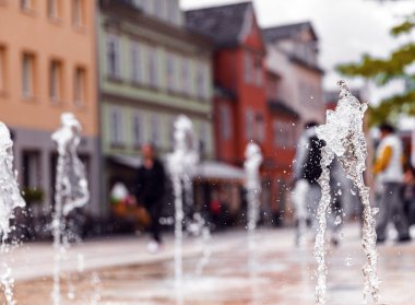 Albertplatz Coburg Bavyera 'daki asfalttan düzinelerce su püskürtüsü fışkırıyor. Arka planda bulanık insanlar ve şehir binalarıyla su sıçratmaya odaklan