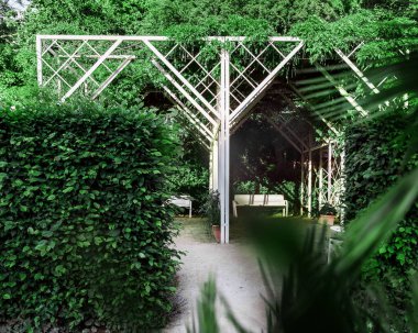 Rosengarten Coburg 'daki beyaz çardak yumuşak odaklı yeşil çit ve palmiye yapraklarından görülebiliyor. Yürüme yolu ve verimli bahçe atmosferiyle Bavyera parkında huzurlu bir yaz günü. Görünürde kimse yok.