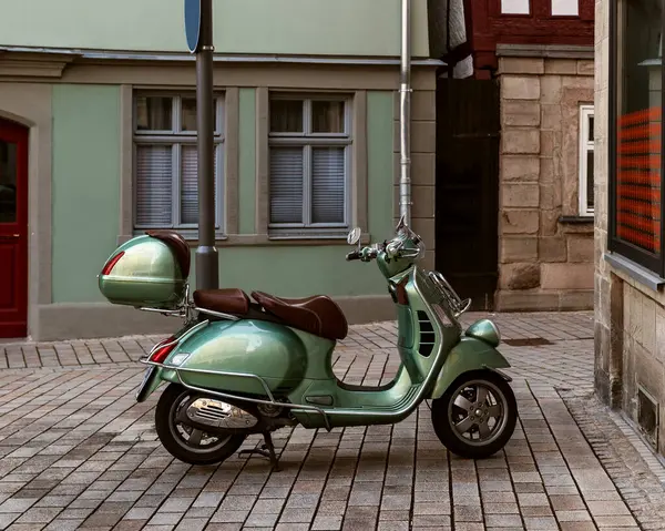Klasik yeşil scooter geleneksel Avrupa evleriyle çevrili sakin bir kaldırım taşı caddesinde park edilmiş. Seyahat, kentsel yaşam tarzı ve klasik hareketlilik temaları için mükemmel bir görüntü.
