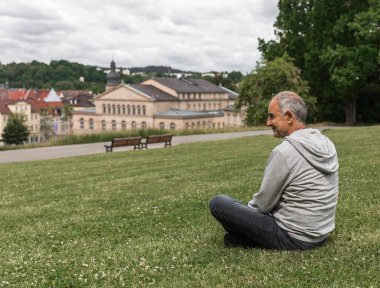 Elderly man in profile sitting near Coburg Hofgarten, panoramic view of historic Ehrenburg Palace and Altes Zeughaus. Reflective moment of solo travel and cultural exploration on sunny day.
