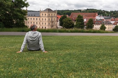 Senior man seen from behind sitting in Coburg Hofgarten, admiring panoramic view of historic Ehrenburg Palace. Peaceful solo travel, active aging, and cultural experience on a sunny day.