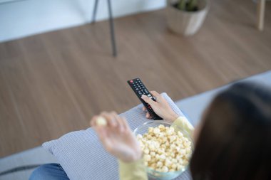 Asyalı bir kadın akıllı televizyon izliyor ve uzaktan kumanda kullanıyor. Uzaktan kumandalı televizyon uzaktan kumandası elinde patlamış mısırla evde.