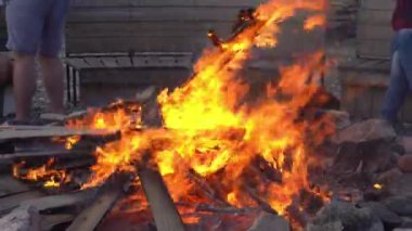 Tanınmayan insanların oturduğu ahşap bankların arka planında şenlik ateşi yanıyor. Yakın planda ateşli bir alev. Parlak renkler ve dinamik alev hareketi. Yüksek kalite 4K görüntü.