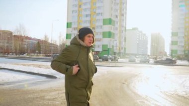 Bir kişi boş bir otoparkta yeşil bir ceket ve spor ayakkabı giyerek dolaşırken, arka planda konut binaları yer alıyor.