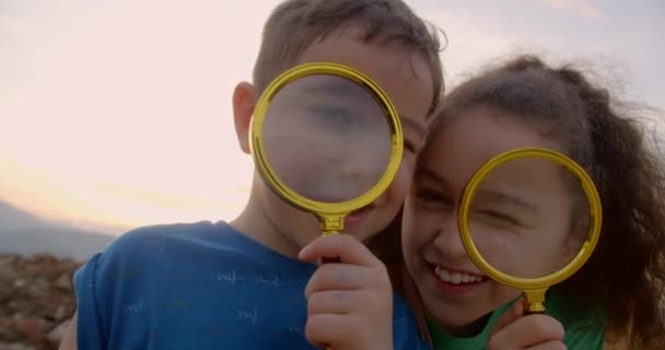 虫眼鏡でカメラを見ている幸せな小さな笑顔の男の子と女の子 公園の芝生の上で子供たちは夢を見ています 幸せなかわいい兄弟姉妹 子供の笑顔とカメラを見上げます — ストック動画