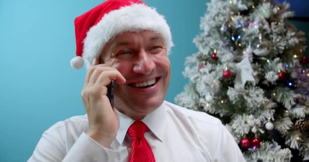 クリスマスツリーの背景に携帯電話を話しているクリスマスハットサンタ クラスの笑顔の男を閉じます — ストック動画