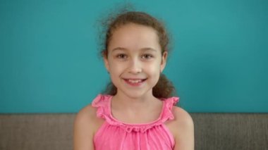 Tatlım, gülümseyen küçük kız evdeki kanepede oturmuş kameraya bakıyor, güzel yüzlü tatlı çocuk kanepede oturuyor. Mutlu bir çocukluk kavramı