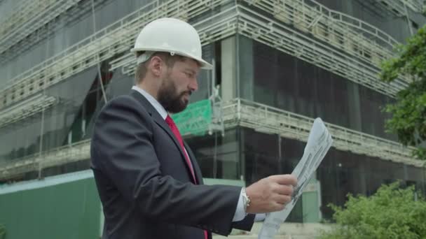 成功した若いハンサムなエンジニア 建築家 建築家 白いヘルメットでビジネスマンの肖像 スーツで 彼の手にプロジェクトを開催し 高層ビルや建設現場の背景 — ストック動画