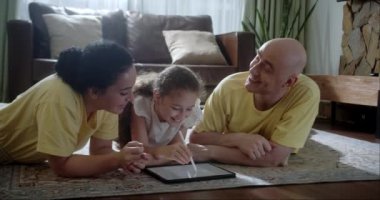 Mutlu aile genç ebeveynleri grafik tablet çizerek tatlı kızın evde sıcak zeminde oturup oyun oynamasını sağlıyorlar anne, baba ve kız boş vakitlerinde oturma odasında eğleniyorlar..