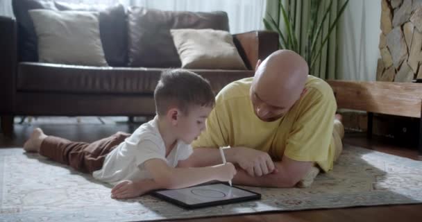 快乐的家庭 年轻的父母们会在平板电脑上画画 帮助可爱的女儿享受躺在温暖地板上聊天的乐趣 爸爸和女孩们也会在客厅里悠闲自在地玩耍 — 图库视频影像